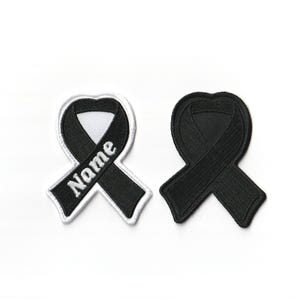 May include: Black ribbon with white stitching and the word "Nome" embroidered on it. The ribbon is in the shape of a heart. There is a second black ribbon in the shape of a heart, but it is not embroidered.
