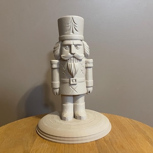 May include: A light-colored wooden nutcracker figurine. The figurine is standing on a round base. The nutcracker is wearing a hat, uniform, and boots. The figurine has a stern expression and a long white beard.