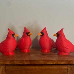 May include: Four red cardinal figurines with black accents around the eyes and orange beaks are displayed on a wooden surface. The birds are positioned in a row, showcasing their vibrant color and detailed design.