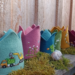 Könnte beinhalten: Ein Set aus sechs Filzkrönchen mit verschiedenen Stickereien. Die Kronen sind in verschiedenen Farben, darunter Grün, Pink, Blau, Gelb und Rot. Die Kronen liegen auf einer mit Moos bedeckten Holzoberfläche.