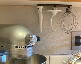 Op de afbeelding: Een zilveren KitchenAid-standmixer met een witte kom en accessoires die aan een rek erboven hangen. De accessoires omvatten een deeghaak, een garde en een platte spatel.