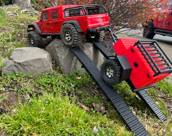 May include: A red toy Jeep pulling a red toy trailer with a black metal cage on top. The Jeep is driving up a black ramp, and the trailer is driving down a black ramp. The Jeep and trailer are on a grassy surface with rocks.