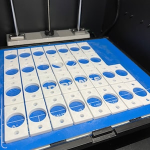 May include: A close-up of a 3D printed bed with a blue surface and a grid pattern.  The bed is filled with white plastic rectangular shapes with circular cutouts.  The text "1. Extruder" is visible on the bed.