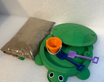 Puede incluir: Un arenero verde con forma de tortuga con tapa, un cubo naranja pequeño y herramientas de juguete azules y moradas. También se incluye una bolsa de arena. El arenero tiene un diseño de cara sonriente.