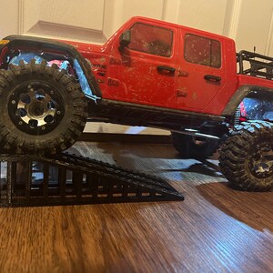 Puede incluir: Una camioneta Jeep Wrangler roja con ruedas negras y una baca negra está subiendo por una rampa de plástico negra. La camioneta está sucia y tiene algunos arañazos en la pintura.