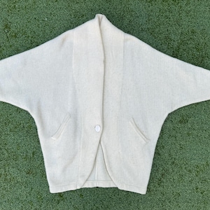 May include: Cream-colored, open-front cardigan with a shawl collar and two front pockets. The sweater has a single button closure and wide sleeves. The fabric appears to be a soft knit. The cardigan is laid flat on a green surface.