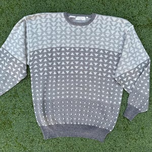 May include: A gray and white patterned sweater with a crew neck. The sweater features a geometric design of triangles and dots, with a gradient effect from light to dark gray. The cuffs and hem are solid gray.
