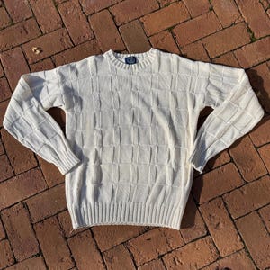 May include: Cream-colored sweater with a textured, grid-like pattern. The long-sleeved knitwear features a ribbed hem and cuffs, and a crew neck. A small label is visible at the neckline. The sweater is made of a soft, comfortable material.