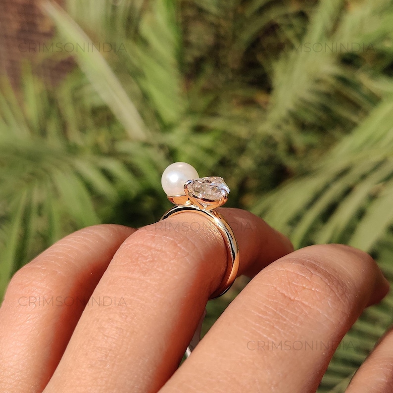 Peut inclure: Une bague en or orn&eacute;e d'une pierre pr&eacute;cieuse claire en forme de c&oelig;ur et d'une perle blanche. La bague est port&eacute;e sur un doigt, avec un arri&egrave;re-plan vert flou.
