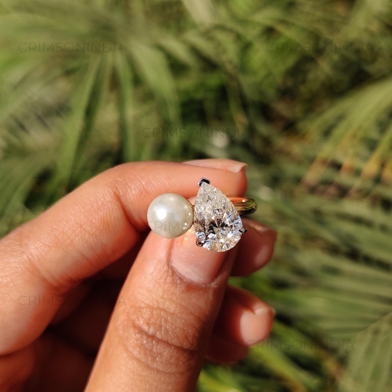 Peut inclure: Une bague en or orn&eacute;e d'un diamant en forme de poire et d'une perle ronde. Le diamant est taill&eacute; &agrave; facettes et la perle est lisse et brillante. L'arri&egrave;re-plan est flou et vert.