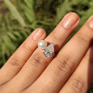 Peut inclure: Une bague en argent orn&eacute;e d'un diamant en forme de poire et d'une perle blanche. Le diamant scintille avec de multiples facettes. La bague est pr&eacute;sent&eacute;e sur un doigt, mettant en valeur le design et l'&eacute;clat du bijou.