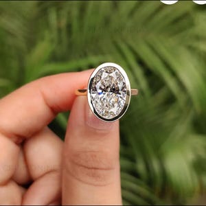 May include: A gold ring featuring a large, oval-cut, clear faceted gemstone set in a smooth, polished gold bezel setting. The ring band is thin and gold, showcasing a clean, minimalist design that highlights the central oval stone's brilliance.