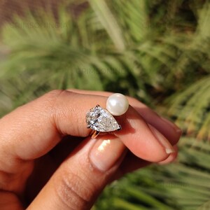 Peut inclure: Une bague en or orn&eacute;e d'un diamant en forme de poire et d'une perle blanche. Le diamant scintille avec de nombreuses facettes, et la perle est ronde et brillante. L'arri&egrave;re-plan est flou et vert.