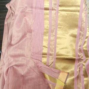 May include: A draped textile in pink and gold. The fabric has vertical stripes and gold borders. The gold sections have a shiny appearance, while the pink sections have a matte finish. The textile is folded, revealing its layered construction. The overall effect is luxurious.
