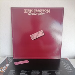 May include: A red vinyl record album cover with the title "Eric Clapton Another Ticket" in white text. A pink ticket with the text "Another Ticket 5008 Eric Clapton" is on the cover.