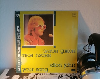Puede incluir: Un disco de vinilo amarillo con una imagen de Elton John usando gafas de sol y una camisa roja. El disco se titula "Your Song" y forma parte de una serie llamada "Archivo de Música Popular".