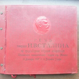 Puede incluir: Un libro rojo con texto en ruso y un retrato de un hombre. El libro está encuadernado con remaches metálicos y tiene una cinta roja. El texto incluye el nombre "И.В.СТАЛИНА" y la fecha "11 Декавра 1937".