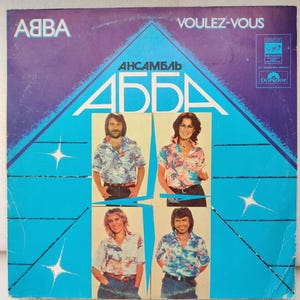 Könnte beinhalten: Ein blaues und weißes Schallplattenalbumcover mit dem Text "Voulez-Vous" und "ABBA" in weißen Buchstaben. Das Albumcover zeigt auch ein Foto der Band ABBA in einem blau-weiß gemusterten Hemd. Das Albumcover trägt den Titel "Ansemble ABBA".
