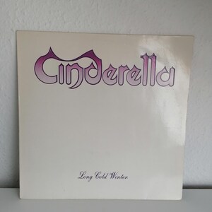 Pode incluir: Uma capa de álbum de vinil branco com o nome da banda "Cinderella" em uma fonte roxa e rosa. O título do álbum "Long Cold Winter" está impresso em uma fonte menor abaixo do nome da banda.