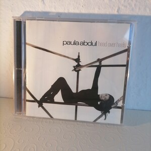 May include: A CD case with a white background and a black and white photo of a woman hanging from a metal structure. The text "paula abdul head over heels" is printed on the case.