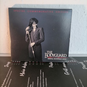 Puede incluir: The Bodyguard Original Soundtrack Album, Edición Conmemorativa Especial. La portada del álbum presenta a una mujer con un traje y un micrófono. El fondo es oscuro, con el título del álbum en blanco.