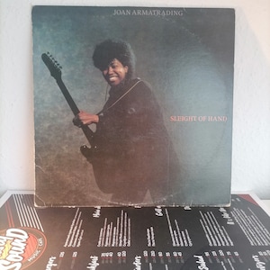 Puede incluir: Portada del álbum "Sleight of Hand" de Joan Armatrading. La portada presenta a una mujer sonriente sosteniendo una guitarra eléctrica negra. El título del álbum está en blanco y el nombre del artista está arriba. El fondo es gris texturizado.