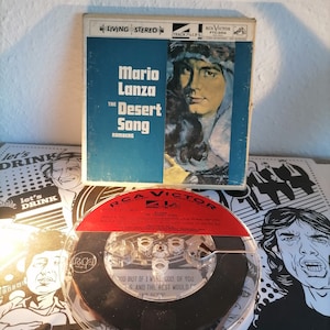 May include: A vintage RCA Victor record album titled "The Desert Song" by Mario Lanza. The album cover is blue and white with a portrait of a man. A clear plastic reel-to-reel tape is in front of the album.