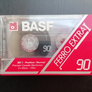 Può includere: Una cassetta audio rossa e argentata con la scritta "BASF Ferro Extra 90". La cassetta è lunga 90 minuti e ha una custodia in plastica trasparente. L'etichetta riporta anche "IEC I - Posizione: Normale Meccanismo di cassetta di precisione 2 x 45min - 135m"
