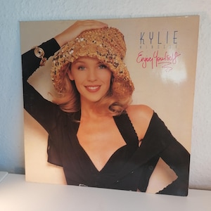 Puede incluir: Una portada de disco de vinilo con Kylie Minogue. La portada es rosa y blanca con una foto de Kylie Minogue con un top negro y un sombrero de lentejuelas doradas. El texto "Kylie Minogue" y "Enjoy Yourself" está impreso en la portada.