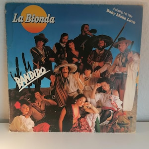 May include: A vintage vinyl record cover featuring a group of people dressed in western attire. The cover is blue and features the text "La Bionda" and "Bandido" in large, bold letters. The text "Including Hit Title Baby Make Love" is also visible in the top right corner.