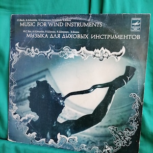 May include: A black and white vinyl record cover with a silver border. The cover features a black and white photo of a statue of a person playing a horn. The text on the cover reads "Music for Wind Instruments" and "Музыка для духовых инструментов".