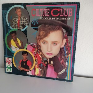 Puede incluir: Una portada de disco de vinilo para el álbum "Colour by Numbers" de Culture Club. La portada presenta una foto de Boy George con una chaqueta rosa, un sombrero negro y una bufanda colorida. El título del álbum está impreso en letras blancas sobre un fondo negro.