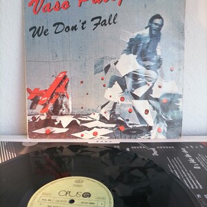 May include: A vinyl record album cover and record featuring a man in a white shirt and blue jeans running through a field of red paper airplanes. The album cover is titled "Vašo Patejdl We Don't Fall" and the record label is "Opus".