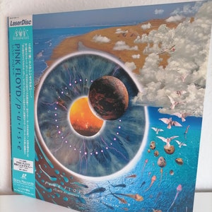 Puede incluir: Un LaserDisc de la película del concierto Pulse de Pink Floyd, que presenta un ojo azul con un planeta rojo y naranja en el centro, rodeado por un anillo de rayos. El fondo muestra un paisaje desértico con nubes y un avión volando por encima.