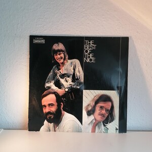 Peut inclure: Une pochette d'album vinyle vintage avec trois hommes. L'album s'intitule "The Best of The Nice" avec le logo Immediate Records. La couverture est principalement noire avec une bordure et du texte blancs. Les hommes sont dans un collage de photos.