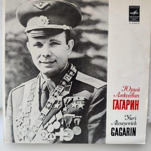 Könnte beinhalten: Ein Schwarzweißfoto von Juri Gagarin, dem ersten Menschen im Weltraum, der eine Militäruniform mit Medaillen trägt. Der Text "Юрий Алексеевич ГАГАРИН" und "Yuri Alexeievich GAGARIN" ist unter dem Foto gedruckt.