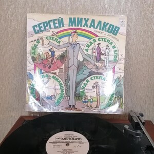 May include: A vintage record cover featuring a cartoon illustration of a man in a blue uniform with a red cap. The cover is titled "Sergei Mikhalkov" and features the text "Dyadia Stepa" in a circle around the man. The cover is in Russian.
