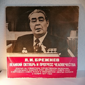 May include: A red and white vinyl record cover featuring a black and white photo of Leonid Brezhnev, a former Soviet leader. The text on the cover reads "L.I. Brezhnev, Great October and the Progress of Humanity, Report at the Joint Solemn Meeting of the Central Committee of the CPSU, the Supreme Soviet of the USSR and the Supreme Soviet of the RSFSR in the Kremlin Palace of Congresses, November 2, 1977."