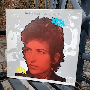 May include: A vintage 3-cassette deluxe edition of Bob Dylan's "Biograph" album. The cover features a close-up of Bob Dylan's face with a red background and a yellow and blue floral design.
