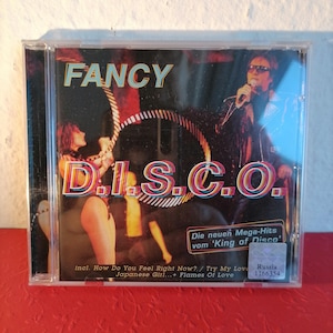 Op de afbeelding: Een CD-hoes met een kleurrijk beeld van een band die op het podium optreedt. De titel van het album is "FANCY D.I.S.C.O." en de tekst "Die neuen Mega-Hits vom 'King of Disco'" is ook zichtbaar. Het album bevat de nummers "How Do You Feel Right Now?", "Try My Love", "Japanese Girl" en "Flames Of Love".