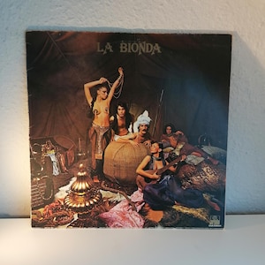 Op de afbeelding: Een vintage vinyl platenhoes met een groep mensen gekleed in kleurrijke, sierlijke kostuums. De albumhoes is getiteld "LA BIONDA" en toont een scène met een vrouw die een snaarinstrument bespeelt.