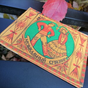May include: A vintage, illustrated card with a red and green design. The card features a man and woman in traditional folk costumes dancing. The text on the card reads "Музыкальный сувенир" which translates to "Musical Souvenir".
