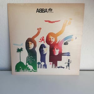 以下が含まれることがあります： カラフルな抽象的なデザインと4つの顔、そして「ABBA The Album」というテキストが書かれた、ビンテージのビニールレコードアルバムのカバーです。