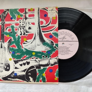 以下が含まれることがあります： 音楽の楽器とクリスマスツリーの枝が描かれた赤いアルバムカバーのヴィンテージビニールレコード。レコードレーベルはピンク色でキリル文字が書かれています。レコードは黒色で、ビニールスリーブに入っています。