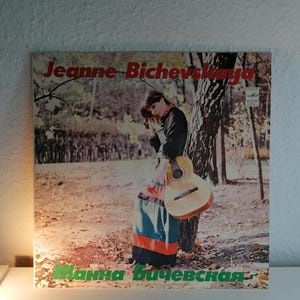 Puede incluir: Una portada de disco de vinilo vintage con una mujer de pelo largo y castaño sosteniendo una guitarra. La portada es de color marrón claro con una falda a rayas verdes y rojas y un árbol en el fondo. El texto de la portada dice "Jeanne Bichevskaya" y "Hanna Bichevskaya".