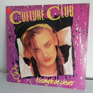 Puede incluir: Una portada de disco de vinilo rosa y negro con una foto de un hombre con el pelo largo y negro que lleva una camisa blanca y un sombrero negro. El texto "CULTURE CLUB" está en letras amarillas en la portada. El texto "kissing to be clever" está en letras amarillas en la parte inferior de la portada.