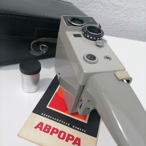 May include: A gray and black vintage 8mm film camera with a leather case and a booklet that reads "КНИГОСЪЕМОЧНАЯ КАМЕРА АВРОРА".