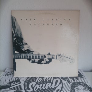 May include: A black and white vinyl record cover with the text "ERIC CLAPTON SLOWHAND" printed in black. The cover features a close-up of a hand holding a guitar.