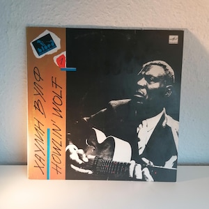 Puede incluir: Una foto en blanco y negro de Howlin' Wolf, un músico de blues, tocando la guitarra. La portada del álbum es naranja y azul con el texto "Howlin' Wolf" y "Kyanah Byrd" en ella.