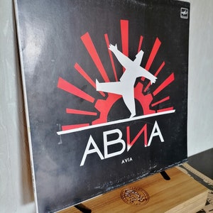 Peut inclure: Un album vinyle vintage avec une couverture noire. Il présente une figure blanche aux bras tendus, des formes géométriques rouges et le mot "АВИА" en blanc. Le mot "AVIA" est en dessous. L'album est présenté sur un présentoir en bois.